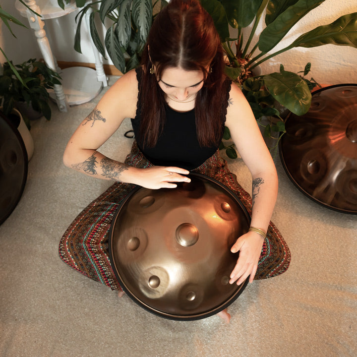 Versierte Handpanspielerin sitzt in einem schönen Raum mit Pflanzen auf dem Boden und spielt eine handgefertigte AeloPan Mini-Handpan mit 9 Tonfeldern aus Edelstahl