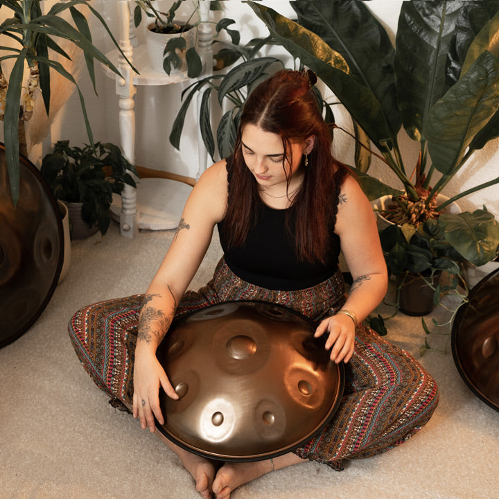 Versierte Handpanspielerin sitzt in einem schönen Raum mit Pflanzen auf dem Boden und spielt eine handgefertigte AeloPan Mini-Handpan mit 9 Tonfeldern aus Edelstahl