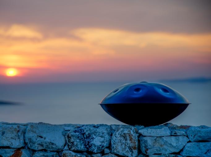 Nitrierte, blauschimmernde Handpan steht auf einer Steinmauer. Im Hintergrund sieht man ein großes Gewässer und einen gelb,rot scheinenden Sonnenuntergang.