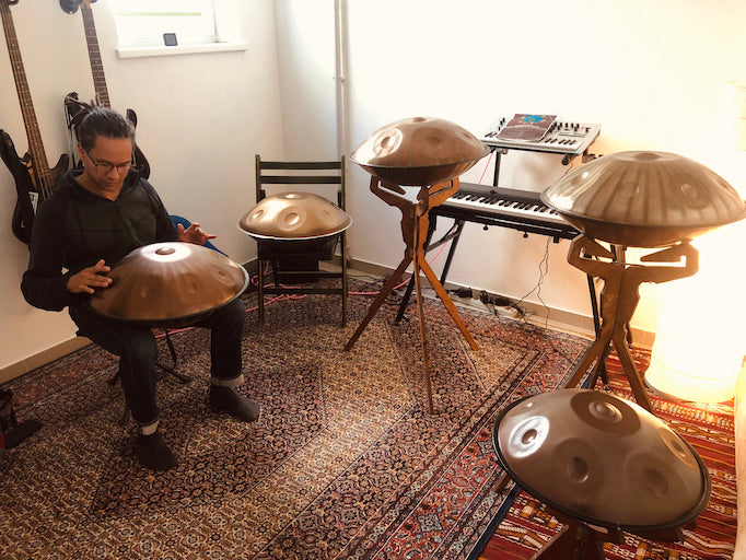 Der Handpan.World Vertreter aus Innsbruck in Österreich sitzt auf einem Stuhl und spielt auf einer Handpan. Zu seiner rechten sind mehrere Handpans auf Handpan Zubehörständer aufgestellt.