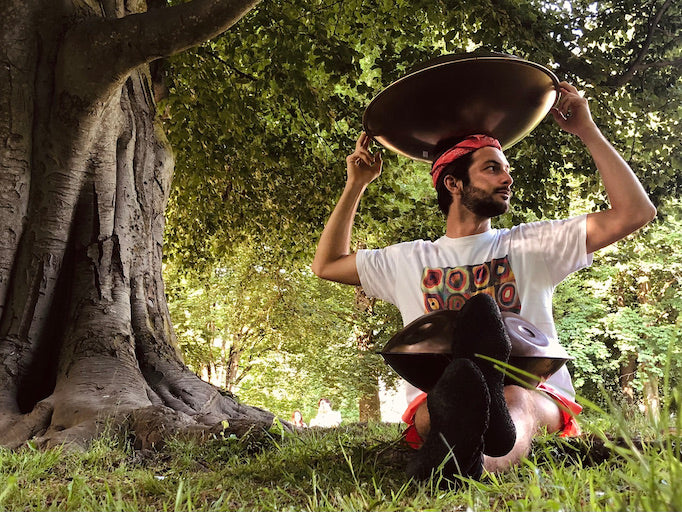 Ein Handpan Spieler sitzt unter einem Baum, auf seinem Schoß liegt eine Handpan und er hält eine Handpan über seinem Kopf.