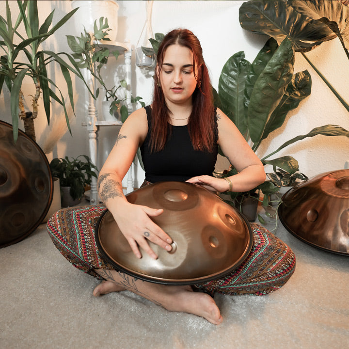 Versierte Handpanspielerin sitzt in einem schönen Raum mit Pflanzen auf dem Boden und spielt eine handgefertigte AeloPan Mini-Handpan mit 9 Tonfeldern aus Edelstahl