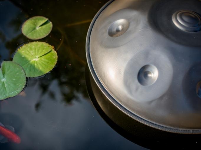 Nitrierte, silberschimmernde Handpan liegt auf einem ruhigen, dunklen Gewässer. Drei Seerosenblätter und ein Goldfisch sind im Wasser zu sehen.