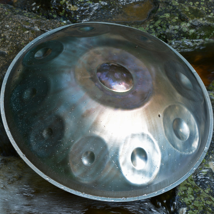  Eine nitrierte Handpan ruht auf einem Felsen neben einem Wasserlauf in einer natürlichen Umgebung.