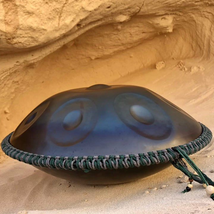 Nitrierte Handpan liegt auf einem sandigen Boden in einer Sandhöhle umrandet mit einem grün-grauen Schmuckband mit hölzernen Perlen am Ende.