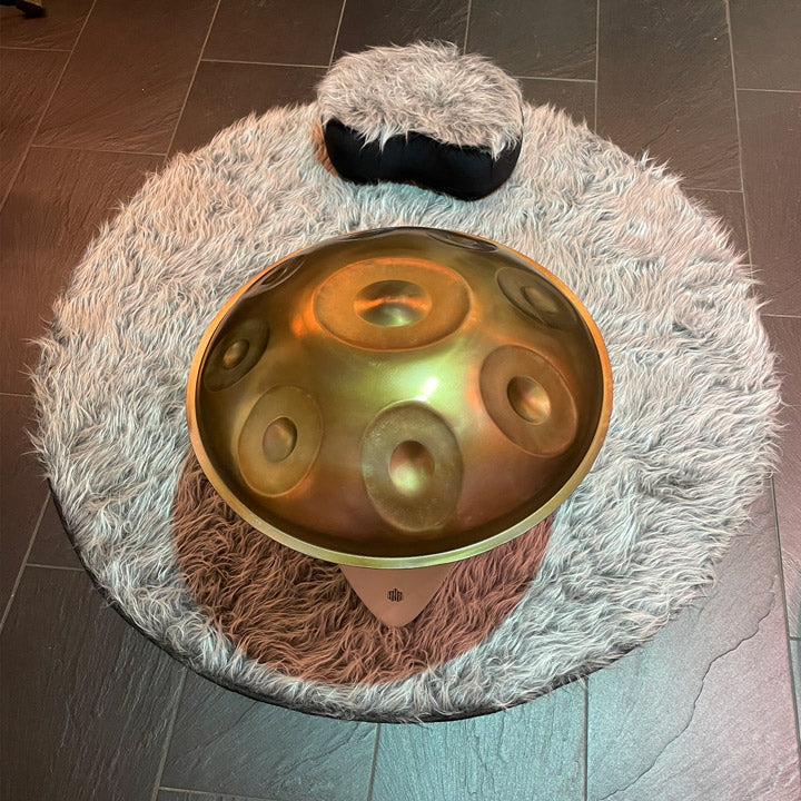 Auf einem praktischen Siddha Handpan Ständer steht eine gold schimmernde Klangskulptur. Der Handpan Halter steht auf einem flauschigen Kissen, davor liegt ein gemütliches Sitzkissen. Das perfekte Set zum Handpan spielen lernen am Boden.