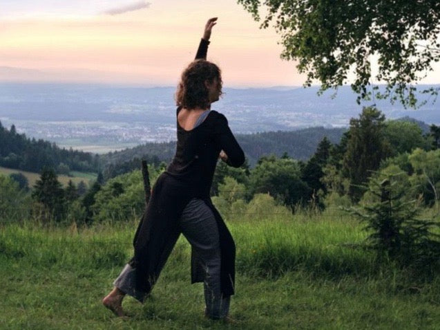 Eine Frau tanzt mit dem Rücken zu uns auf einem Hügel im Grünen bei Sonnenaufgangs Stimmung. Im Hintergrund sieht man das Tal