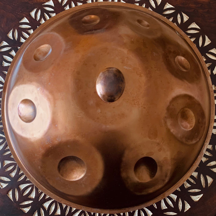 Bronzefarbene Embersteel Svaraa Handpan Drum von oben mit neun Tonfeldern liegt auf einem hölzernen Boden mit Muster. Diese Handpan, sowie viele weitere kann man online bei Handpan.World sicher erwerben.