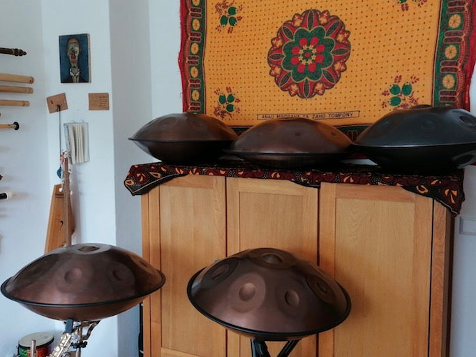 Zwei Handpans aus Edelstahl stehen im Vordergrund auf Ständer und drei weitere liegen im Hintergrund auf einem kleinen Schrank.