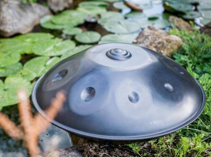 Silberschimmernde, nitrierte Handpan liegt am Rande eines Baches, welcher mit Seerosenblättern bedeckt ist. Am Rand des Baches liegen große Steine.