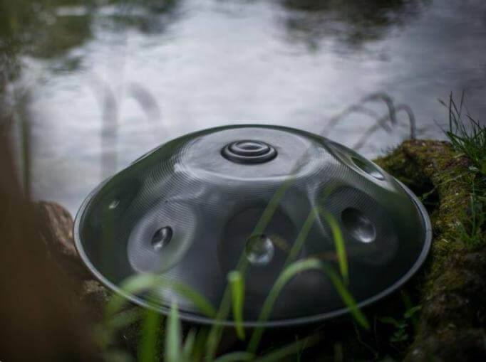 Nitrierte, silberschimmernde Handpan liegt am Rande eines Gewässers, welches den grau-weißen Himmel widerspiegelt. Im Vordergrund ragen grüne Stränge hervor, die die Handpan leicht verdecken.