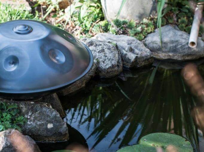 Links sieht man eine nitrierte Handpan mit leichtem blau-silbernen Schimmer. Davor liegt ein Bach der von großen Steinen umrandet ist und ein Rohr, dass in den Bach führt.