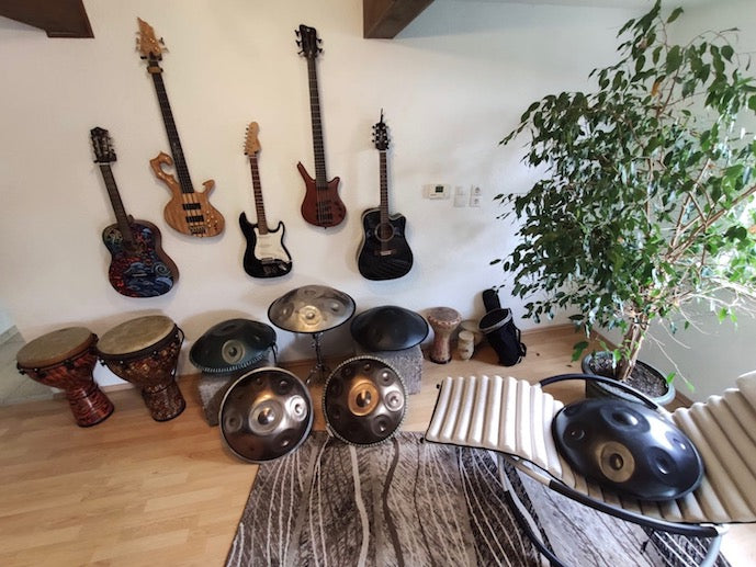 Im Handpan Showroom Mannheim hängen vier E-Gitarren an einer Wand, fünf Handpans und zwei Trommeln sind vor der Wand aufgestellt. In der Ecke steht ein Fikus