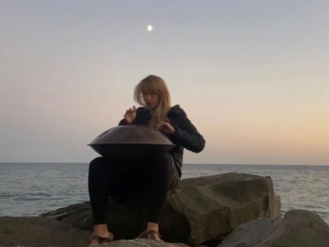 Jenny Geilke sitzt in der Abenddämmerung auf einem Felsen am Meer und spielt Handpan