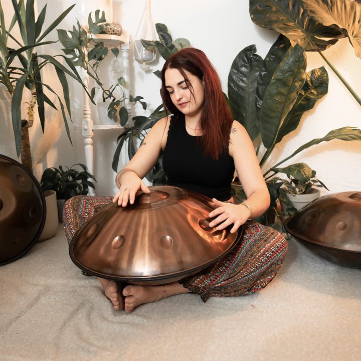 Handpanspielerin sitzt in einem schönen Raum mit Pflanzen und Handpans auf dem Boden und spielt eine AeloPan Handpan, die Bronze glänzt