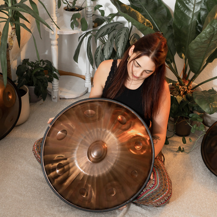 Handpanspielerin zeigt die handgefertigte, Bronze schimmernde AeloPan Handpan. Sie sitzt in einem gemütlichen Raum, umgeben von Pflanzen und Handpans auf dem Boden