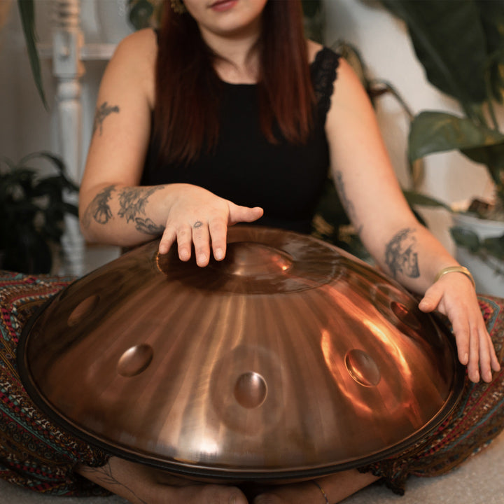 Nahaufnahme einer AeloPan Handpan auf dem Schoß einer versierten Handpanspielerin. Die Handpan schimmert Bronze gestreift. Im hintergrund sind Pflanzen zu sehen