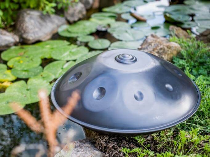 Nitrierte, silberschimmernde Handpan liegt an einem Bachufer, welcher fast komplett mit Seerosenblättern bedeckt ist. 