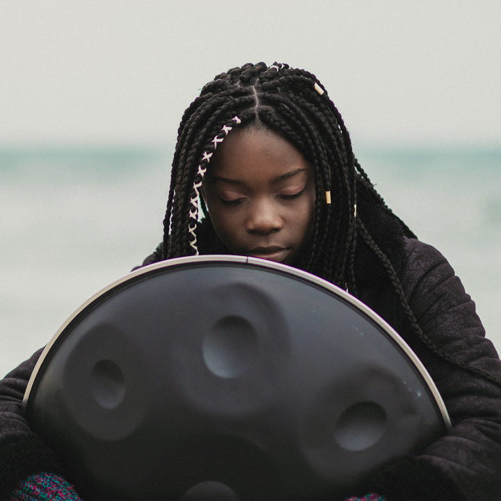 Frau hält eine nitrierte Black Magic Handpan in den Armen. Im Hintergrund ist ein Gewässer zu sehen.