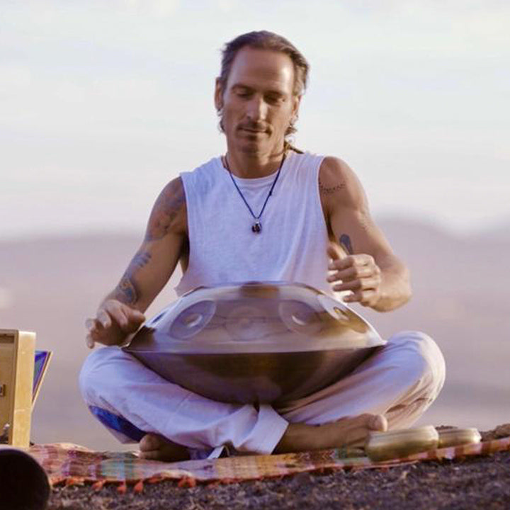 Handpanspieler beim spielen auf einer NamiPan. Er sitzt in Mitten von großen Bergen und trägt weiße Klamotten.
