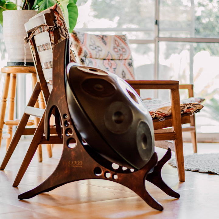 Ein hölzerner Dekorationsständer für Handpans steht in mitten eines lebendigen Wohnzimmers mit gemütlichen Stühlen und Pflanzen im Hintergrund. Auf dem Ständer steht eine nitrierte Handpan mit einem silbernen Schimmer.