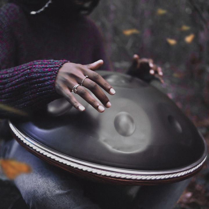 Handpanspielerin spielt auf einer nitrierten Handpan mit leichtem silbernen Schimmer. Sie sitzt in Mitten eines dunklen Waldes, während sie von gelben Schmetterlingen umflogen wird.