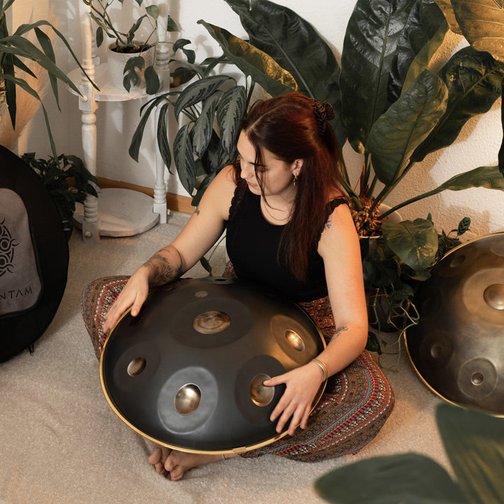 Versierte Handpanspielerin sitzt in einem schönen Raum mit Pflanzen auf dem Boden und spielt eine handgefertigte KitaPantam Handpan 432Hz mit 9 Tonfeldern in dem Design Polished Gold