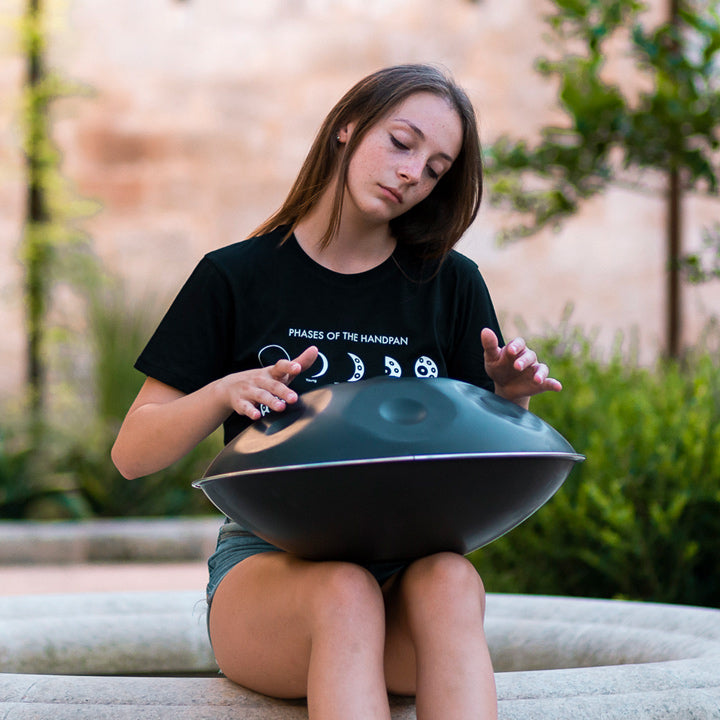 Eine Frau hat eine nitrierte Handpan auf dem Schoß und spielt darauf, während sie auf einem Steinbrunnenrand sitzt. Im Hintergrund steht eine Backsteinmauer und viele grüne Pflanzen sind zu sehen.