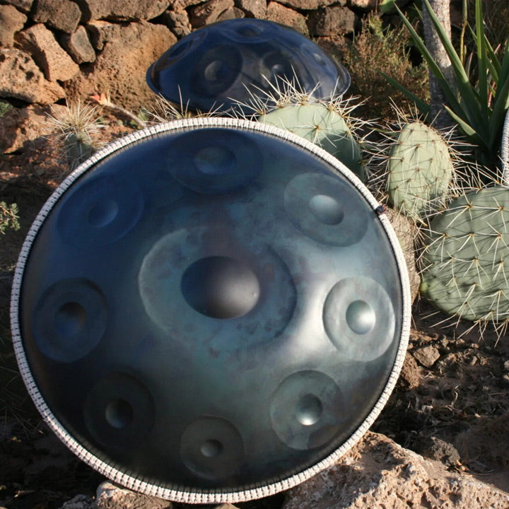 Drei nitrierte Handpans stehen auf erdig-sandigen Boden zwischen einem Kaktus und Palmen im Hintergrund. Sie schimmern leicht Silber und sind umrandet mit weißen Schmuckbändern.
