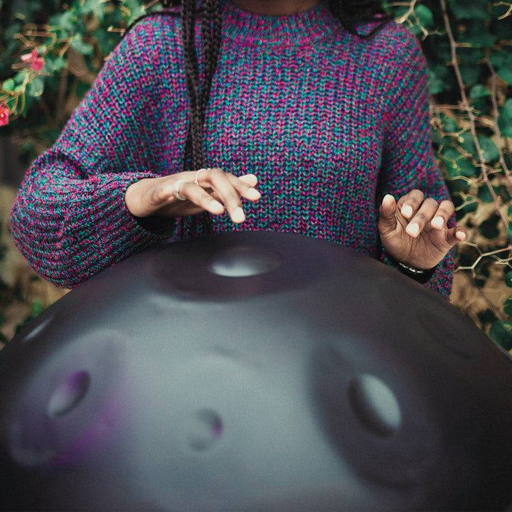 Eine Frau mit einem lila-grün-blauem Strickpullover spielt auf einer dunkelgrauen, nitrierten Handpan. Im Hintergrund ist ein Rosenbusch mit rosa-roten Blüten und grünen Blättern.