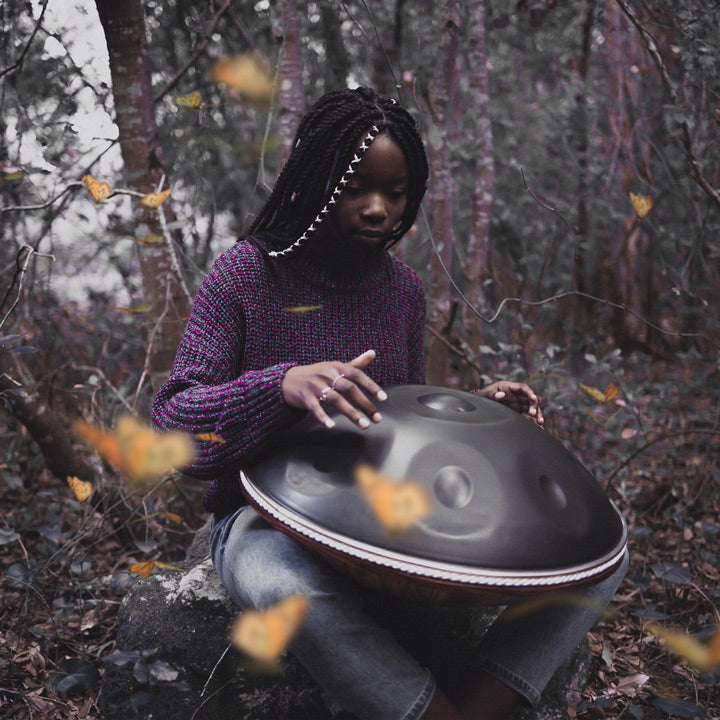 Eine Frau spielt auf einer nitrierten, dunkelgrauen Handpan in der Mitte eines dunkelgrünen und brauen Waldes, während gelbe Schmetterlinge um sie kreisen.