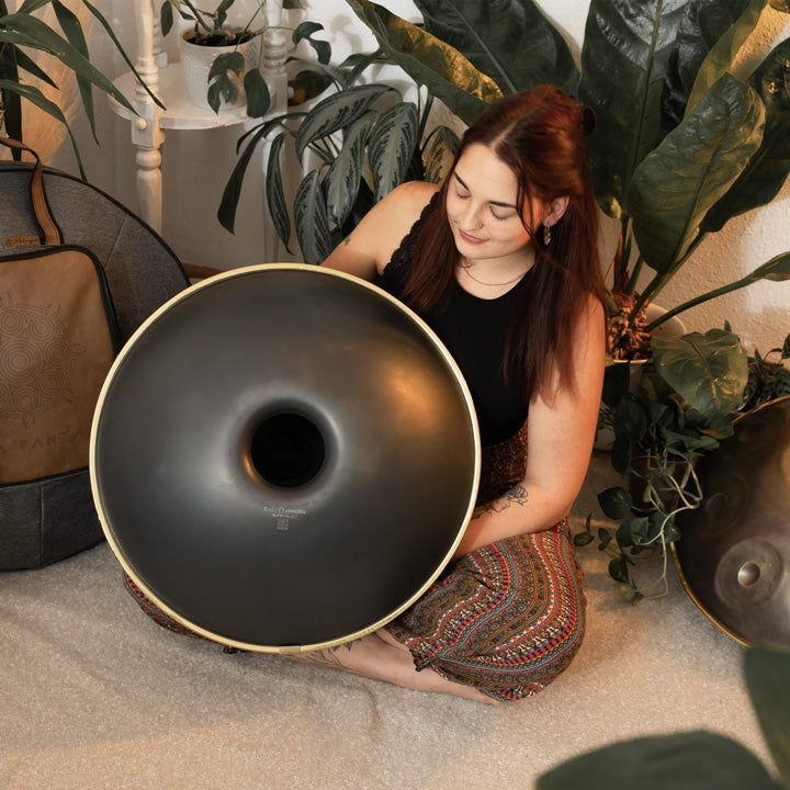 Handpanspielerin zeigt die schwarze Unterseite einer KitaPantam Handpan aus nitriertem Stahl. Sie sitzt auf einem Teppich zwischen Pflanzen und Handpans