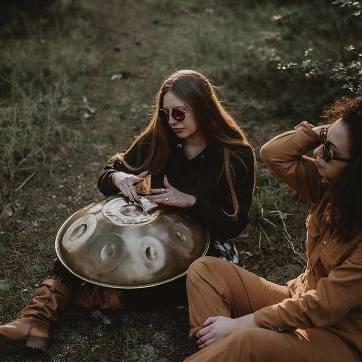 Zwei Frauen sitzen auf einer Wiese, während eine auf einer goldenen, silberschimmernden Handpan spielt und die andere den wundervollen Klängen lauscht. Diese Handpan, sowie viele weitere kann man online im Handpan.World Store kaufen.