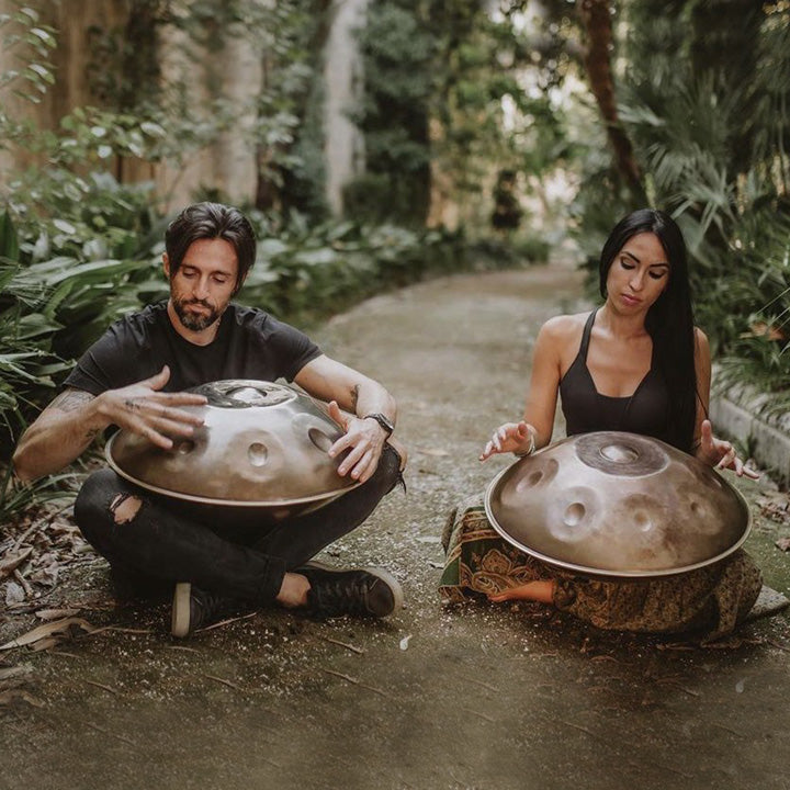 Zwei Handpanspieler spielen auf goldenen silberschimmernden Handpans und sitzen dabei auf einem erdigen Weg umrandet von grünen Pflanzen. Diese Handpan, sowie viele weitere kann man online im Handpan.World Store kaufen.