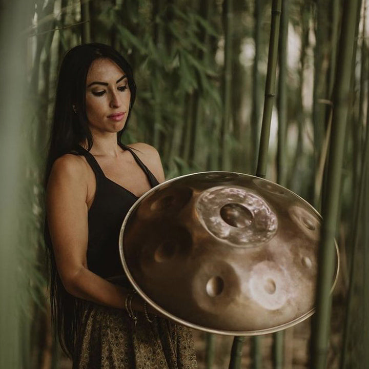 Eine Handpanspielerin hält eine goldene Handpan in den Händen während sie zwischen mehreren Bambussträngen lehnt. Die Handpan spiegelt sich im Licht und schimmert dadurch silbern. Diese Handpan, sowie viele weitere kann man online im Handpan.World Store k