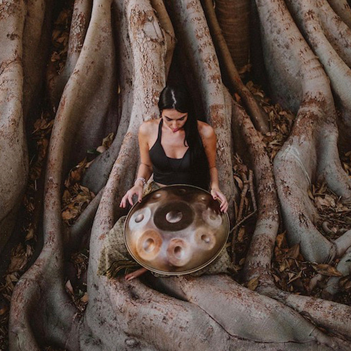 Eine Handpanspielerin spielt eine Spiritwave Handpan am Fuße eines größen Baumes. Diese uns weitere Handpans findest Du bei Handpan.World