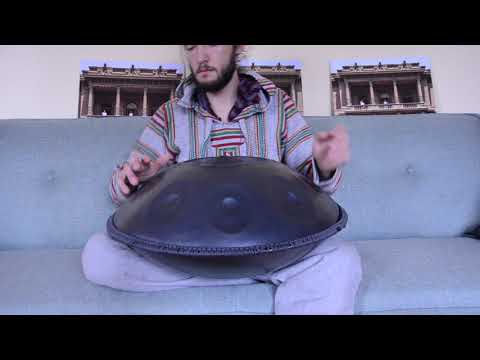 Handpanspieler spielt auf einer C# Kurd Handpan mit einem grauen Schmuckband während er auf einer grauen Couch in einem hellen Wohnzimmer sitzt.