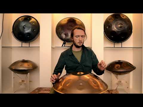 Braunhaariger Handpanspieler spielt auf einer E Sabye Handpan. Um ihn herum stehen fünf weitere Handpans in einem weißen Regal. Diese Handpan, sowie viele weitere kann man online im Handpan.World Store kaufen.