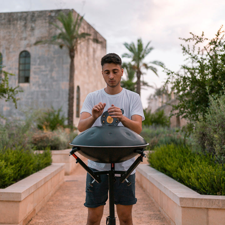 Ein Mann spielt auf einer dunkelgrauen, nitrierten Handpan, die auf einem hohen, schwarzen Ständer steht. Hinter ihm sind grüne Pflanzen, Palmen und ein Steinhaus.